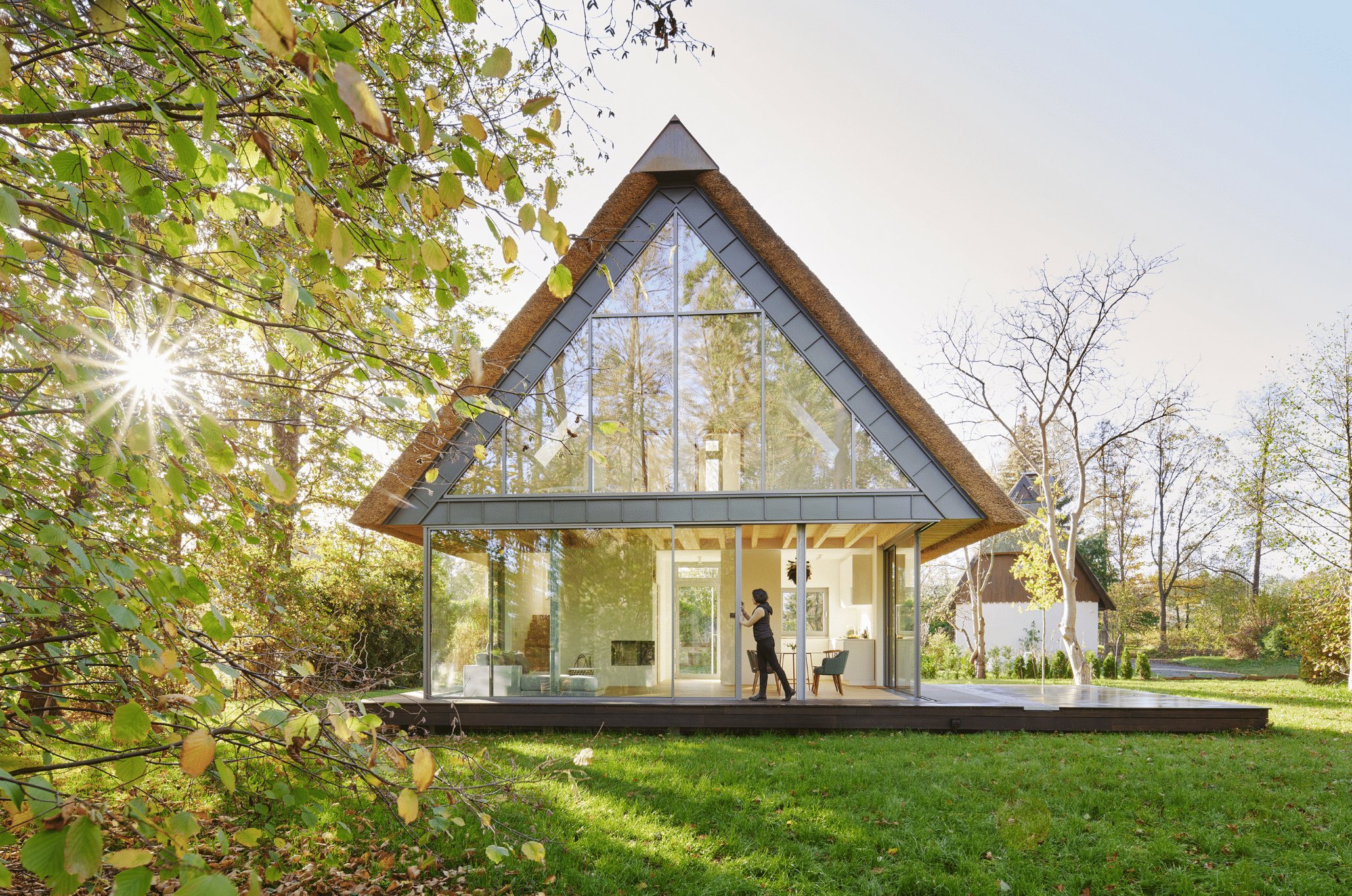 Solarlux zeigt ein Tiny House mit einer Glasschiebetür und grossen Glasfronten mit einer Terrasse und spitzem Dach in einem gepflegten Garten.