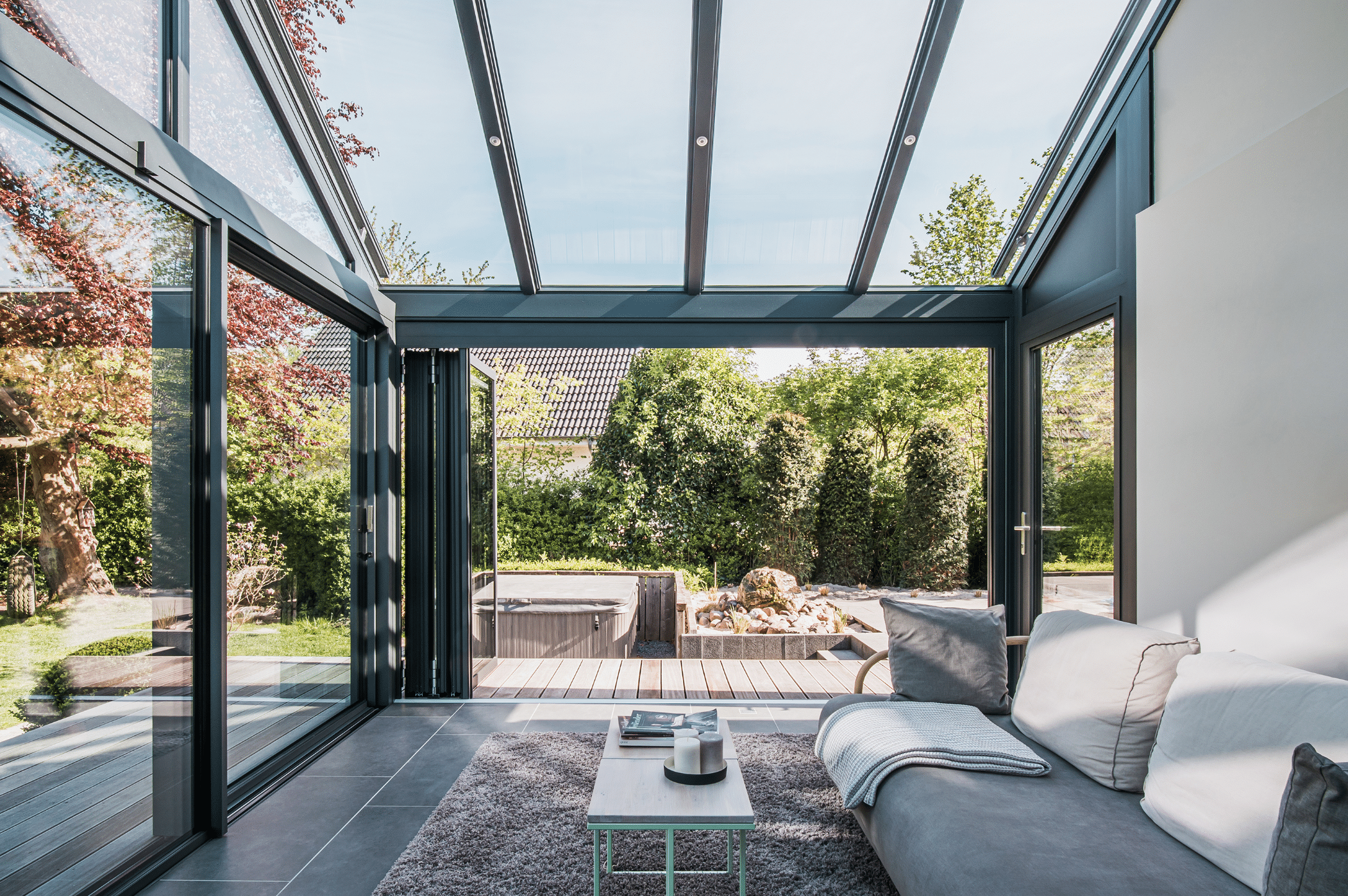 Solarlux zeigt einen gläsernen Wintergarten mit Glasschiebetüren und einem Panoramadach mit gemütlicher Sitzlandschaft und freiliegendem Teppich.