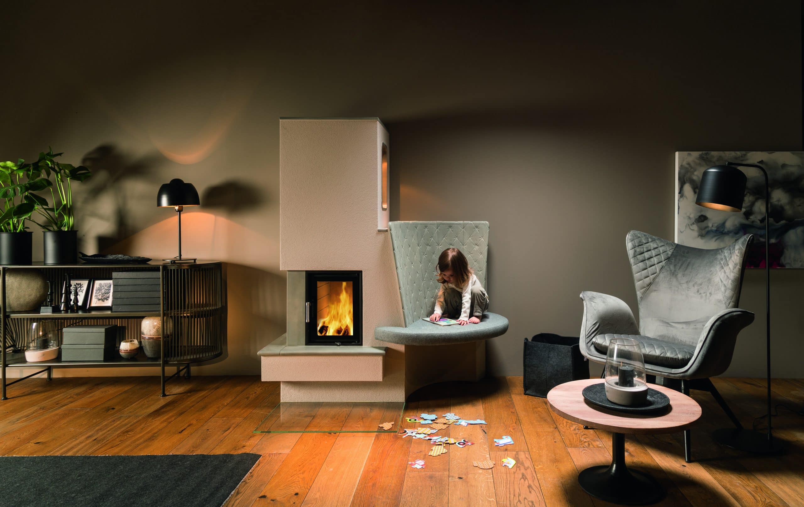Sommerhuber zeigt ein Wohnzimmer mit weissem Kachelofen mit Sitzmöglichkeit und einen silbergrauen Ohrensessel mit einem Beistelltisch aus Holz.