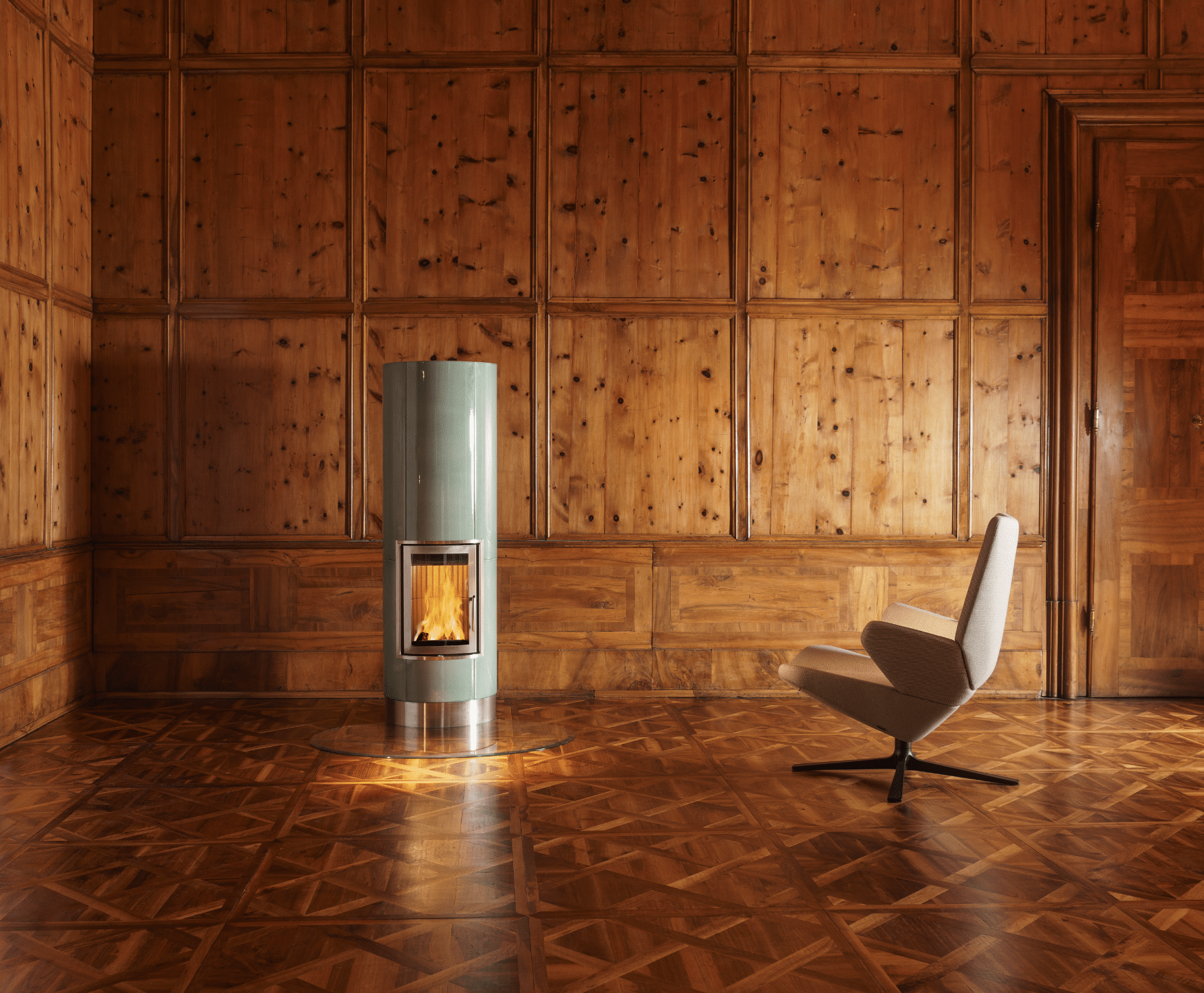 Sommerhuber zeigt einen Raum mit Holzvertäfelung an allen Wänden und gemustertem Holzboden mit einem Vollspeicherkachelofen in blassgrün und silber.