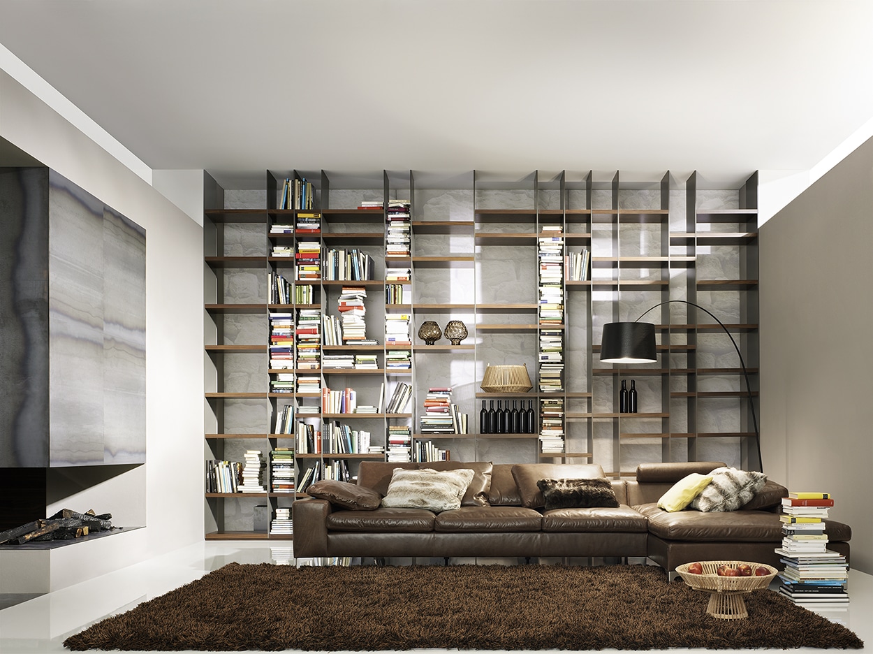 Mit Möbeln von haas ausgestattetes Wohnzimmer im Stil einer Bibliothek mit hohem, offenem Bücherregal, brauner Ledercouch, Kamin und Teppich.