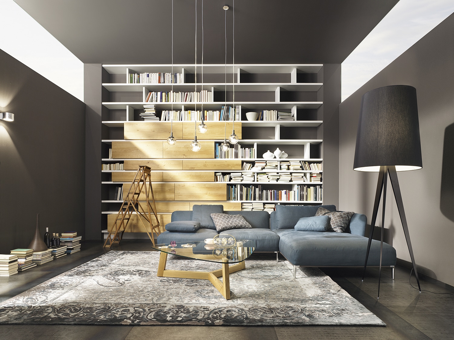 Mit Möbeln von haas ausgestattetes Wohnzimmer im Stil einer Bibliothek mit hohem, teils offenem Bücherregal samt Leiter, blauer Couch, auffälliger Stehlampe und Teppich.