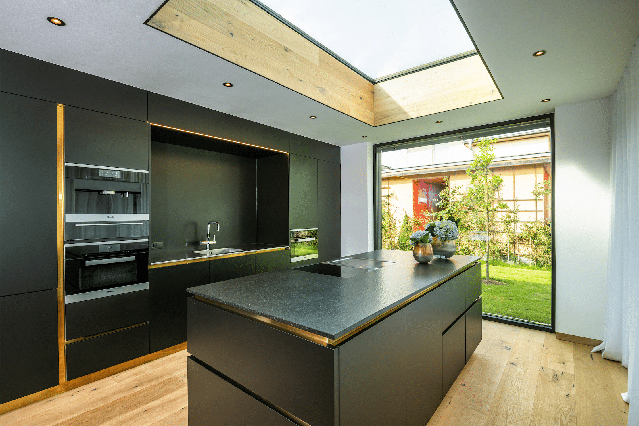 Moderne Einbauküche mit Kücheninsel in schwarz mit deckenhohem Fenster und Dachlichte sowie Holzboden von der Tischlerei Oberreiter.