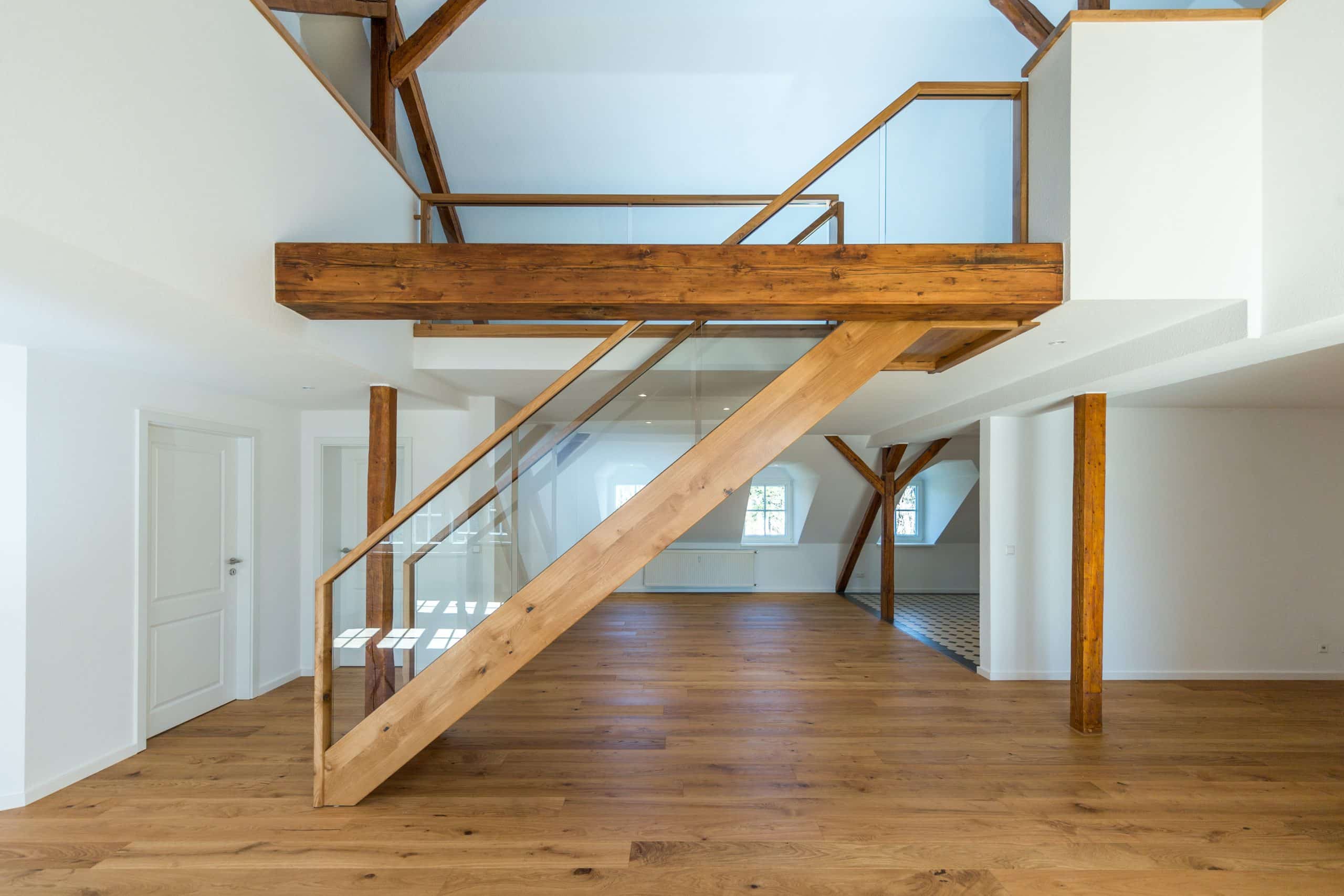 Treppenmeister präsentiert einen modernen Treppenaufgang ins Dachgeschoß, in einem großen lichtdurchfluteten Raum mit Glasgeländer und dazu passendem Holzboden.