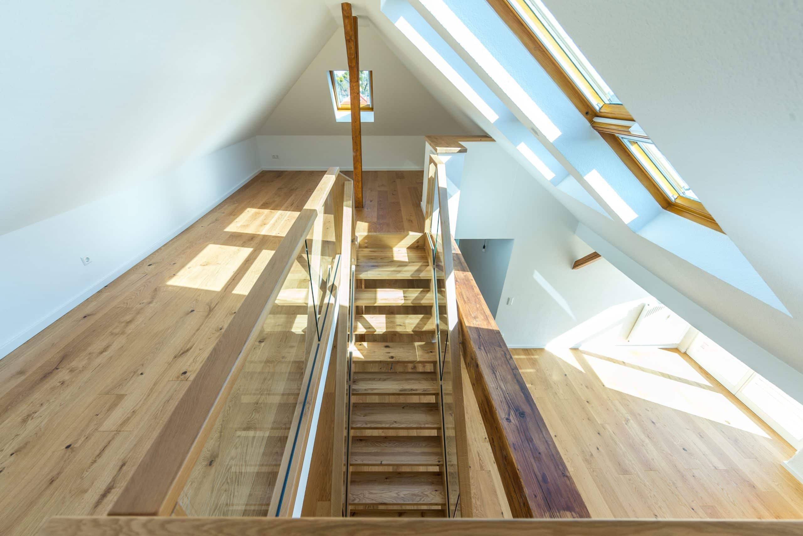 Treppenmeister präsentiert einen modernen Treppenaufgang ins Dachgeschoß, in einem großen lichtdurchfluteten Raum mit Glasgeländer und dazu passendem Holzboden.