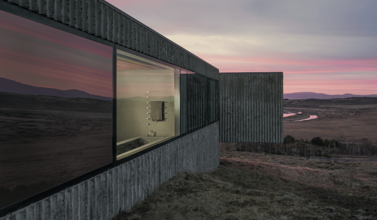 VOLA zeigt ein Betonhaus von außen mit großem Fenster und Blick in das Badezimmer mit Badewanne, silbernen Armaturen und Handtuchhalter an der Wand.