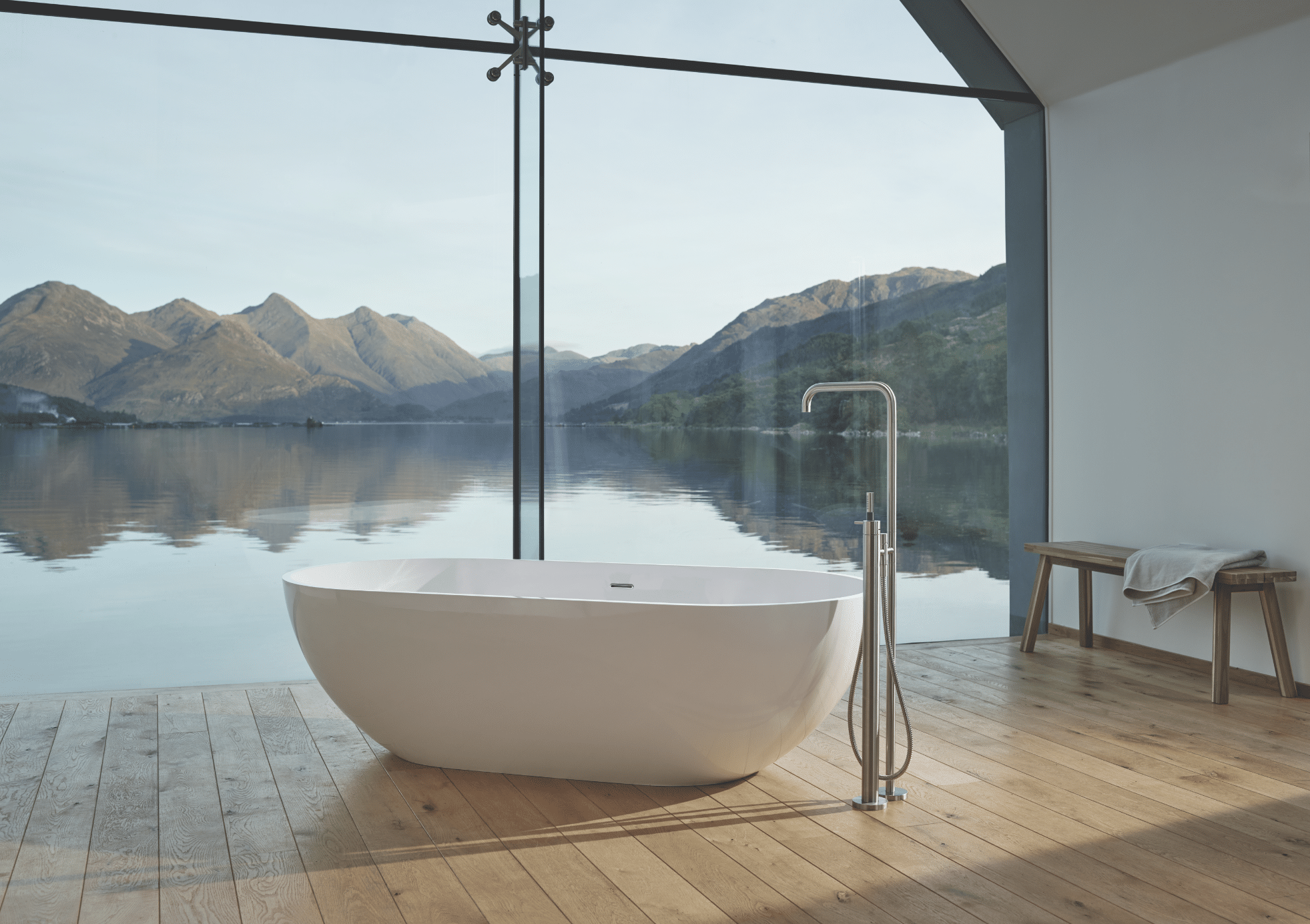 VOLA zeigt ein Badezimmer mit Holzboden und einer weissen, freistehenden Badewanne mit silbernen Armaturen und großer Glasfront mit Blick auf einen See.