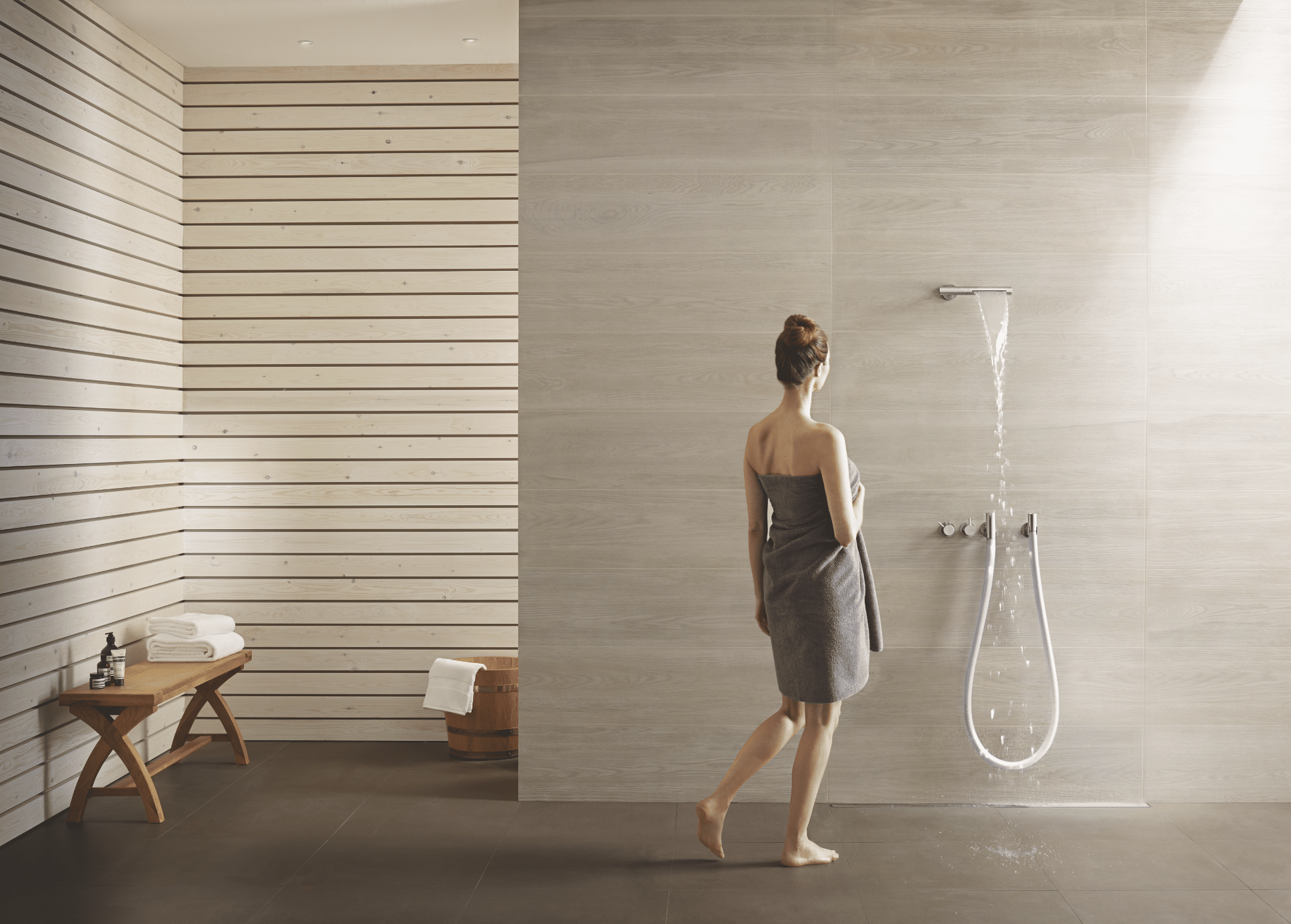 VOLA zeigt ein offenes Badezimmer mit Fliesenboden, Holzwänden und mit ebenerdiger Dusche und silbernen Armaturen.