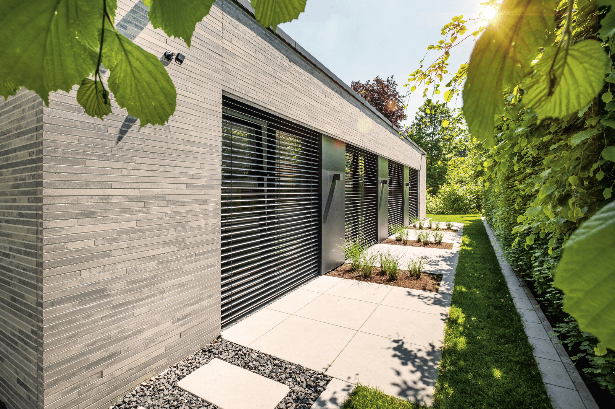 WAREMA zeigt eine Seitenwand eines hellen Hauses mit großen Glasfronten, dunklen Jalousien und einem Weg aus Steinplatten mit Pflanzen zu dem Garten.