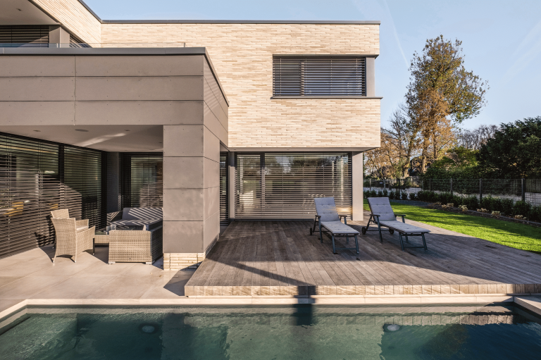 WAREMA präsentiert ein helles Haus mit einer überdachten Terrasse, dunklen Jalousien, einer Essgruppe aus Rattanmöbeln und eine Sonnenterrasse aus Holz mit Liegen neben einem Pool.