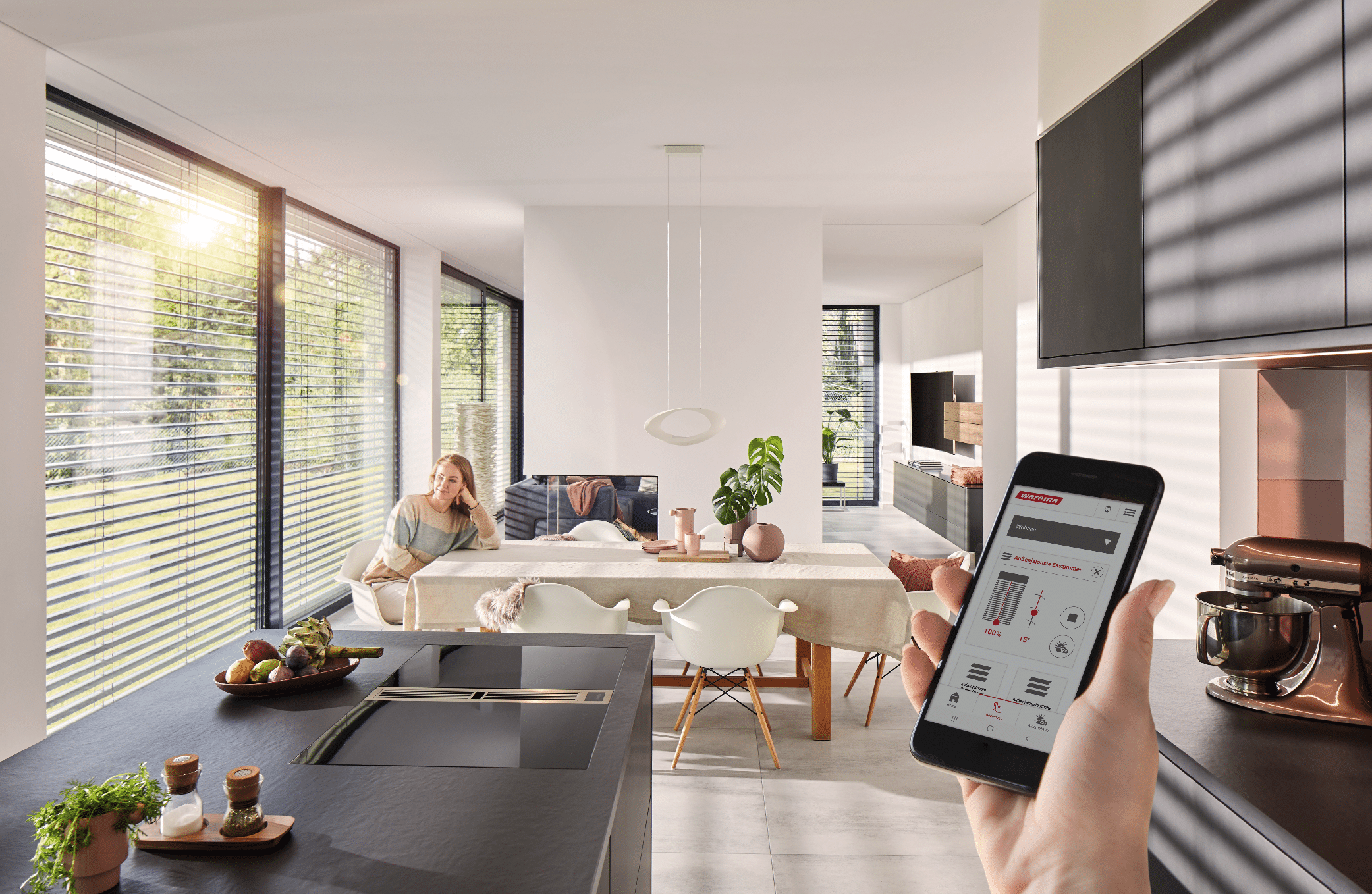 WAREMA zeigt einen schwarzen Küchenblock mit Blick auf das Esszimmer und großen Fenstern mit dunklen Außenjalousien, die über das Handy steuerbar sind.