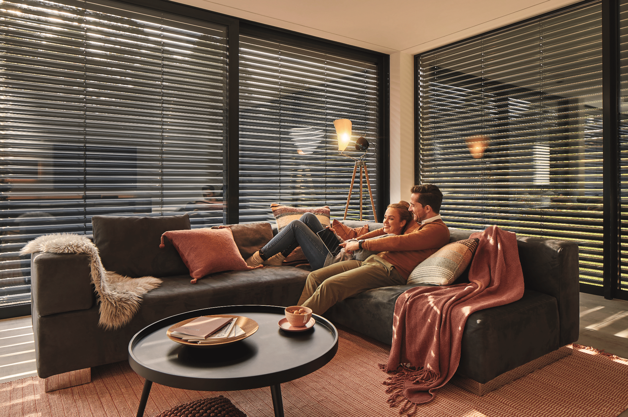 WAREMA zeigt ein mit Jalousien abgedunkeltes Wohnzimmer, eine braune Couch mit verschiedenen Polstern und einem runden Beistelltisch aus Holz.