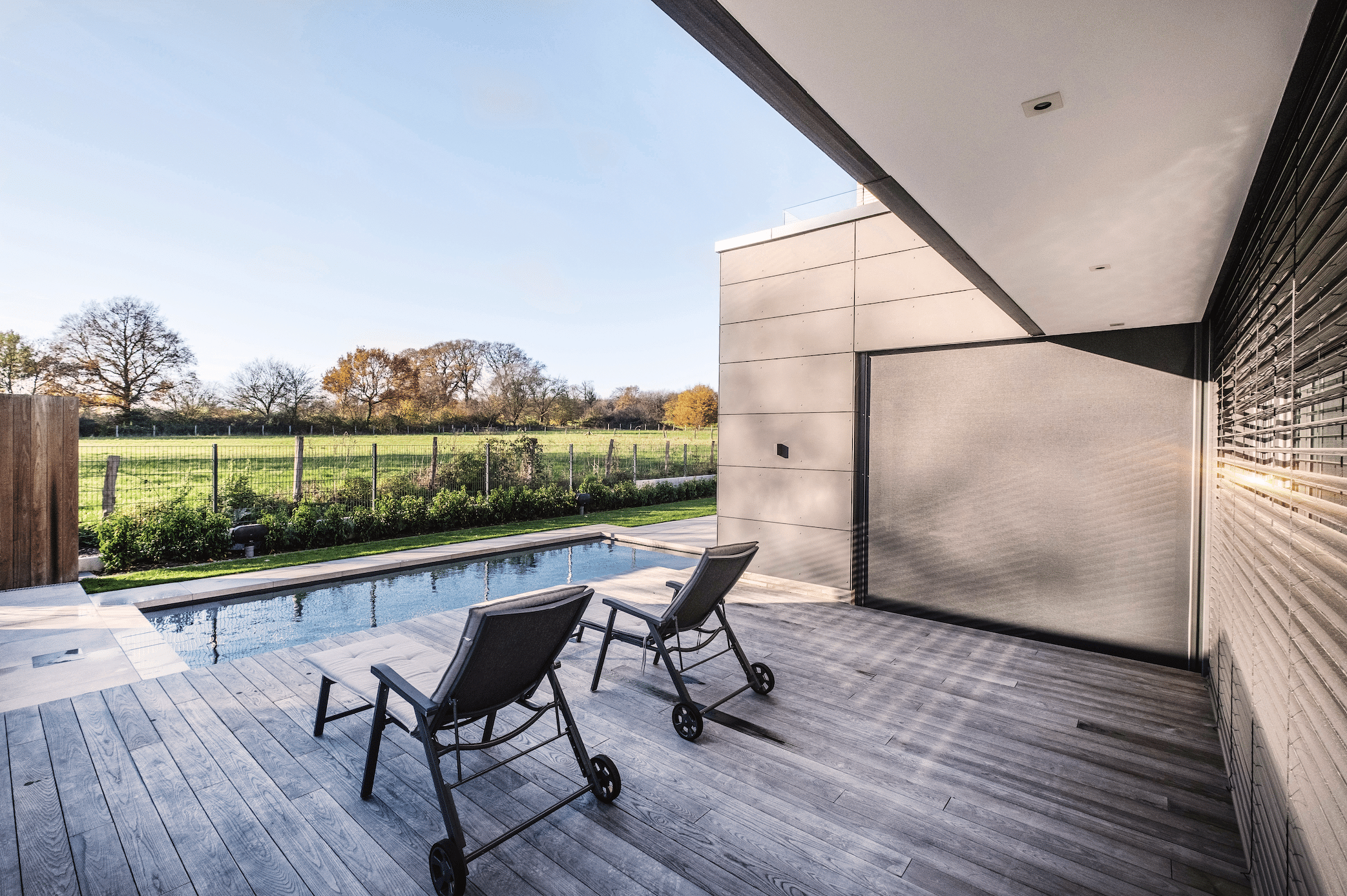 WAREMA präsentiert einen Außenbereich mit Pool und einer Sonnenterrasse mit Holzdielen und Liegen vor einer mit Jalousien abgedunkelten Terrassentüre.