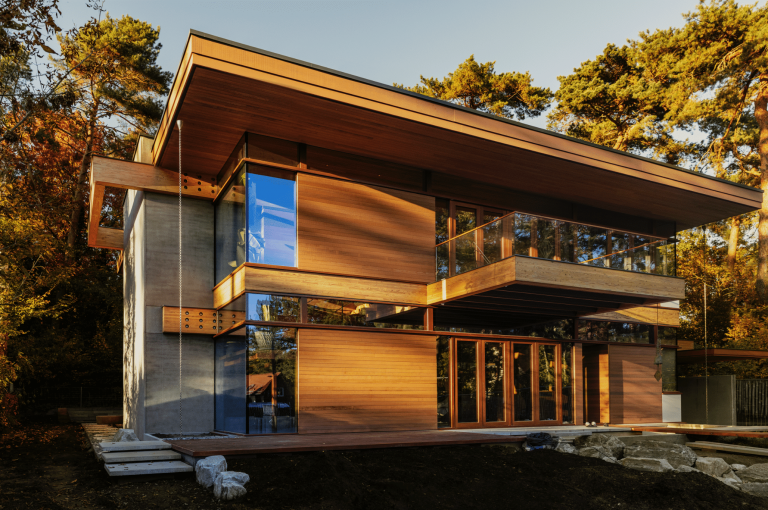 Weissenseer Holz-System-Bau zeigt ein schönes Einfamilienhaus aus Holz mit großem Balkon, vielen hohen Fenstern und überdachter Terrasse im Grünen.