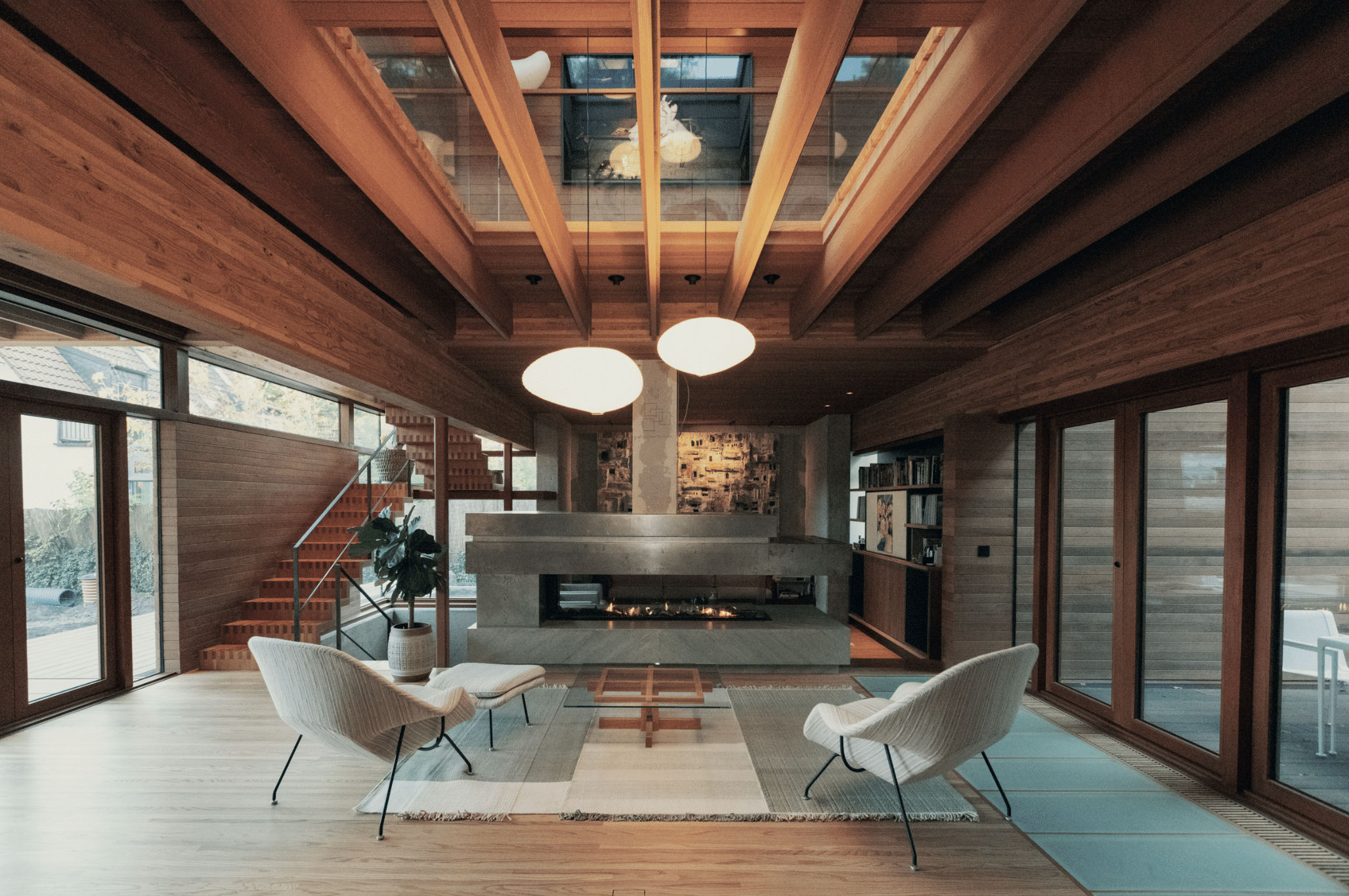 Weissenseer Holz-System-Bau zeigt ein gemütliches Wohnzimmer in einem Holzhaus mit Loungesesseln, passenden Hockern und freiliegendem Teppich und mit Blick auf einen modernen Kamin.