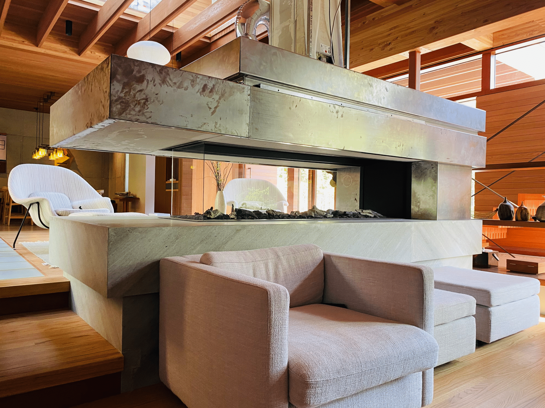 Weissenseer Holz-System-Bau zeigt ein gemütliches Wohnzimmer in einem Holzhaus mit einem Kamin aus Stein und gemütlichem Sessel in einem hellen Pastellton davor und passenden Hockern.
