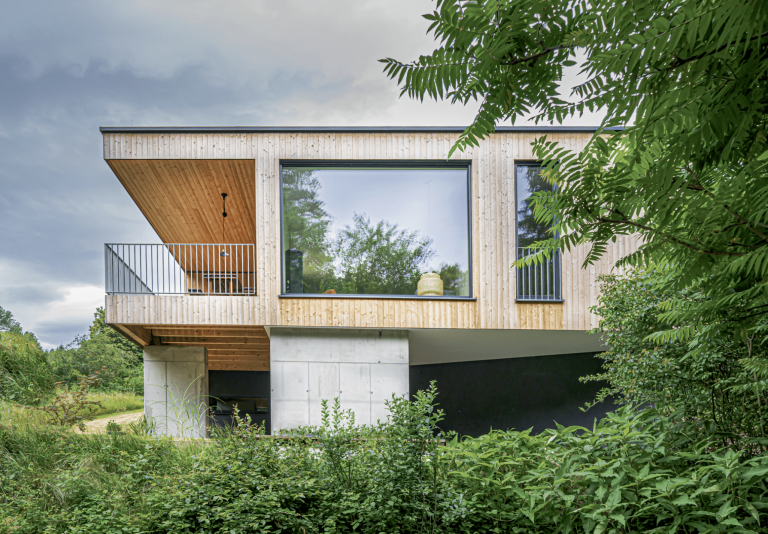 Weissenseer Holz-System-Bau zeigt modernes Flachdach-Wohnhaus mit Holzriegelfassade auf Betonstehern, nach innen versetztem Balkon und großer Glasfront.