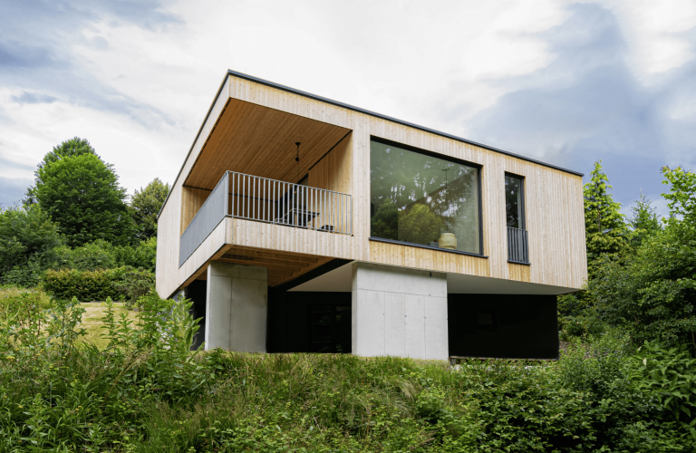 Weissenseer Holz-System-Bau zeigt modernes Flachdach-Wohnhaus mit Holzriegelfassade auf Betonstehern, nach innen versetztem Balkon und großer Glasfront.