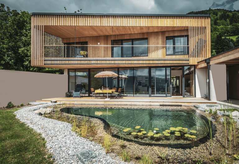 Modernes Einfamilienhaus mit Flachdach, Holzfassade, Balkon überdachter Terrasse und Schwimmteich von Weissenseer Holz-System-Bau.