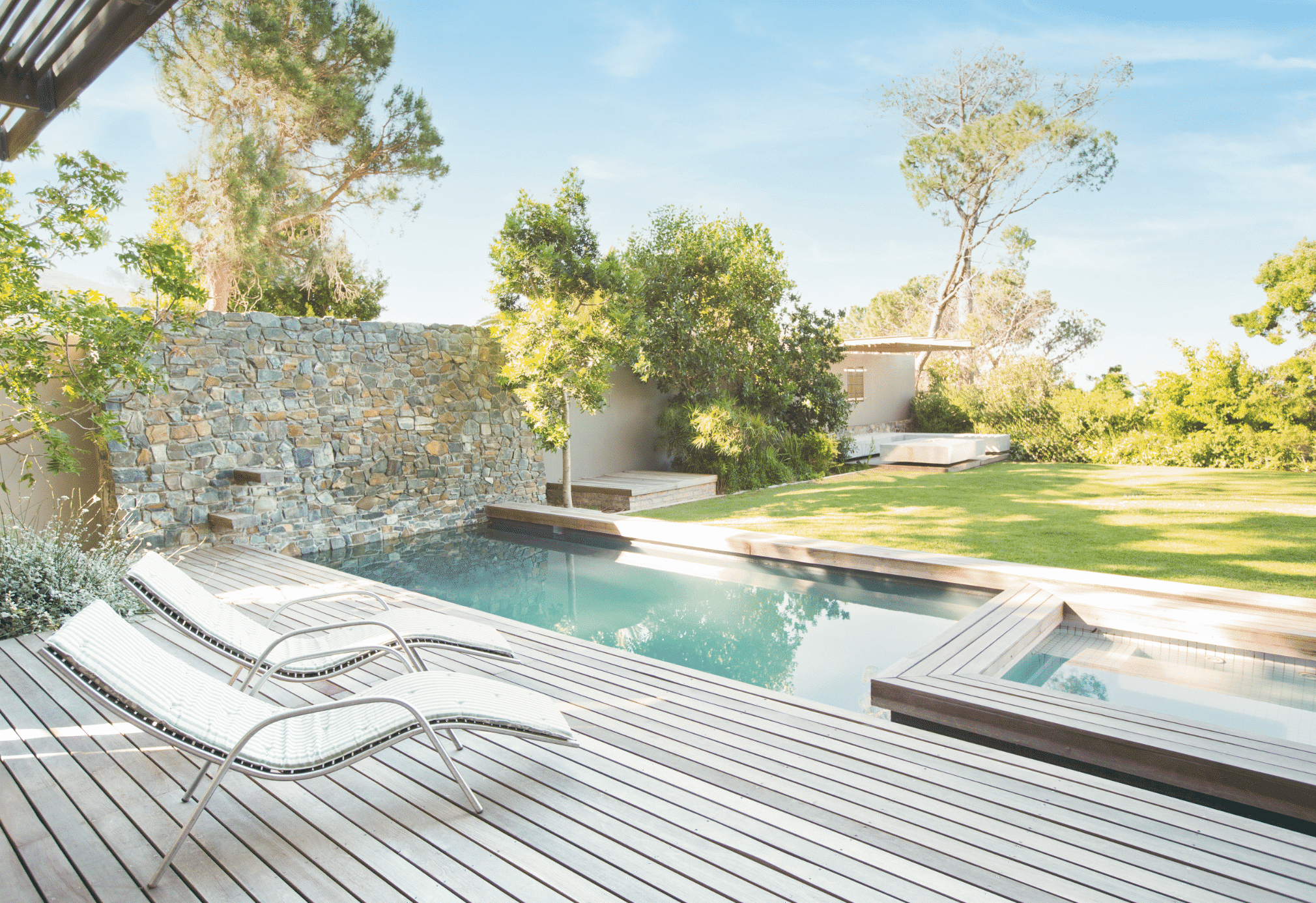 Moderne Poolanlage mit Natursteinwand und grauer Terrassen und Poolumrandung aus Holz im großem Garten und Baumbestand.