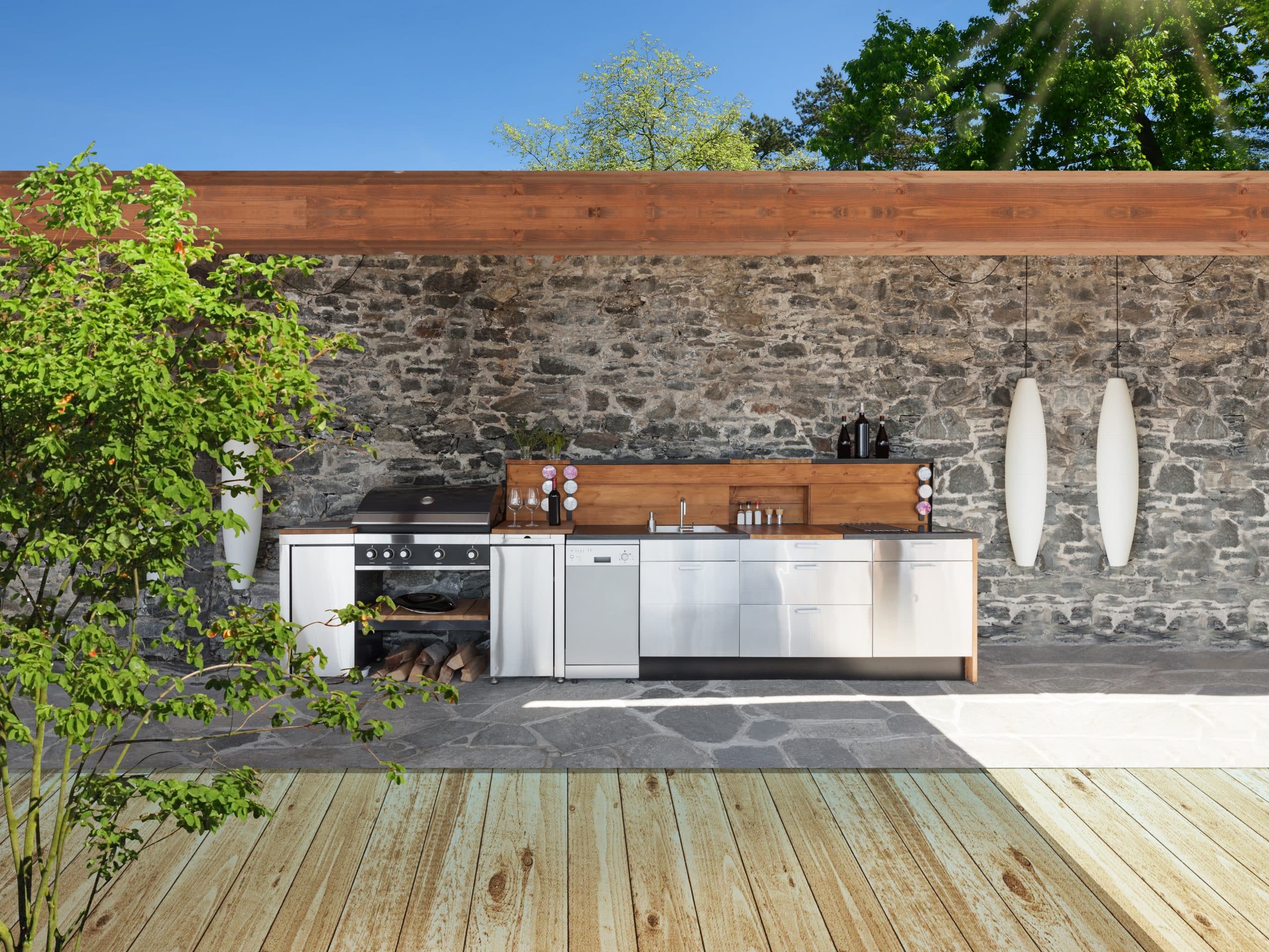 Moderne Outdoorküche mit Elementen aus Holz und Nirosta-Edelstahl überdacht und witterungsgeschützt an einer Natursteinwand.
